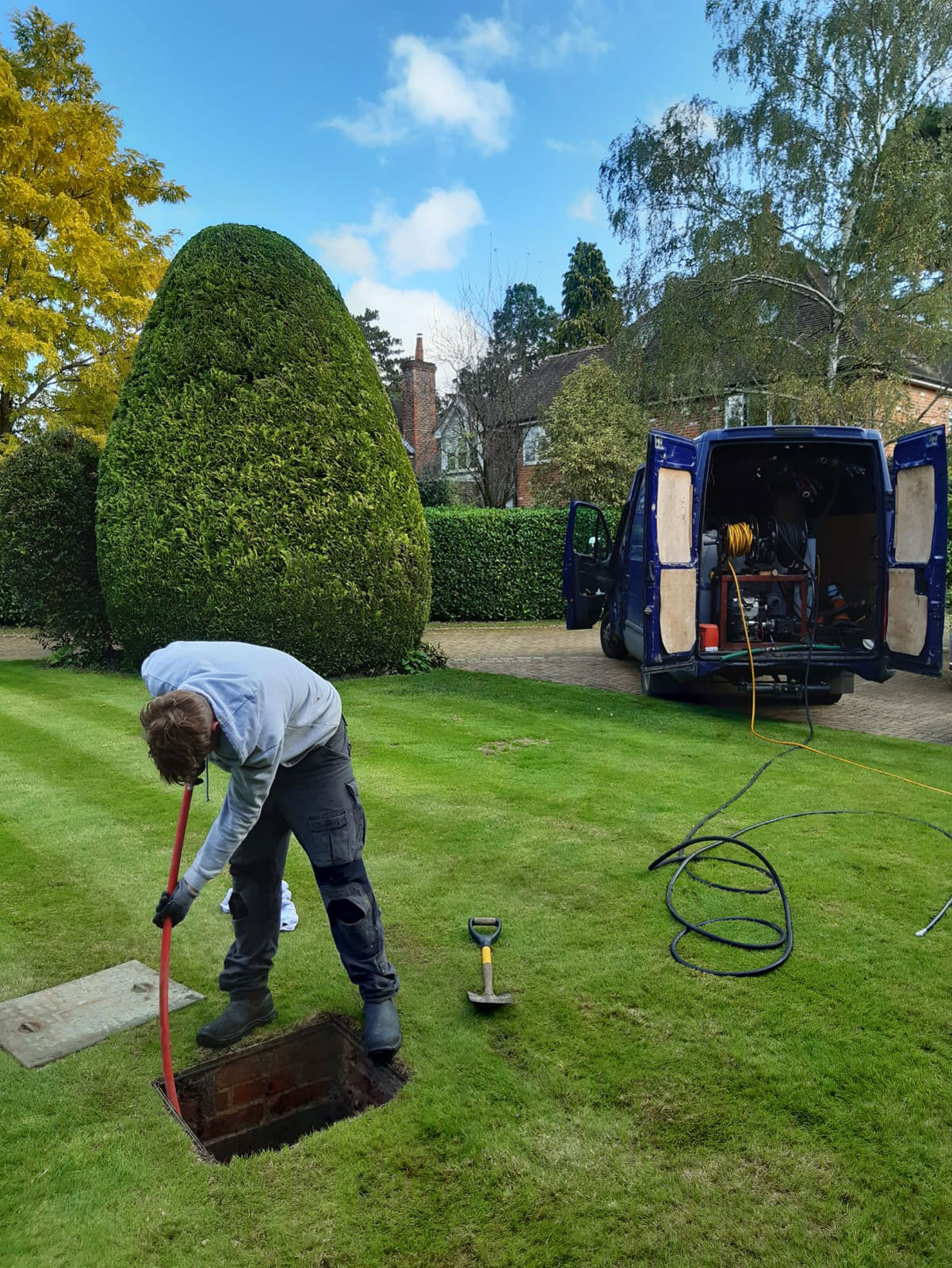 Declan from Drain Ace jet washing a blocked drain in Fairmile Lane Cobham with the manhole open on a landscaped lawn